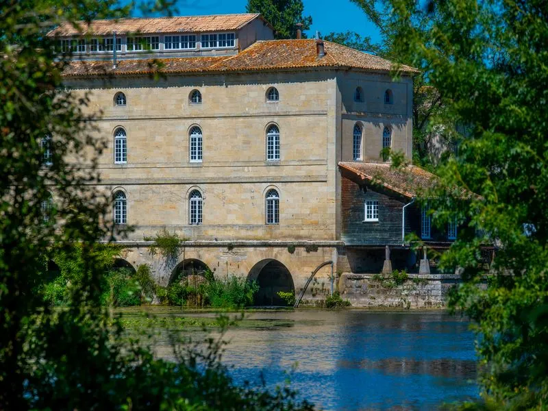 Porchères Mill in Bordeaux, an 18th-century stone building beside the Garonne River, featuring regular facades with arched windows and a stone arch spanning the water, surrounded by lush green vegetation.