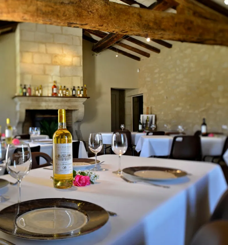 Elegant dining room at Château Haut-Bergeron in Bordeaux featuring set tables, wine glasses, a bottle of white wine, fireplace, and exposed wooden beams.