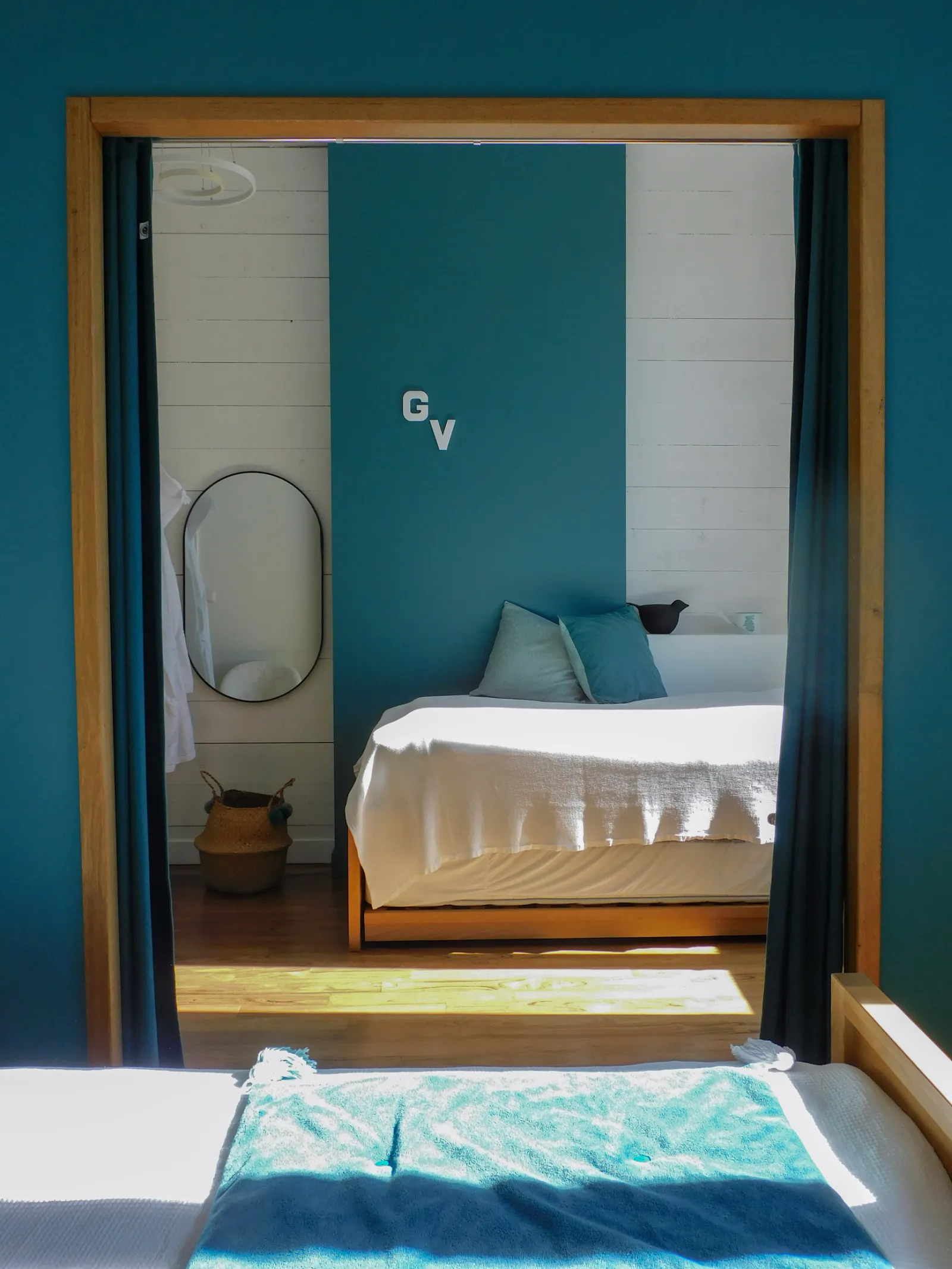 Modern hotel room with teal walls, wooden bed with white linens and blue pillows, oval mirror, and ensuite bathroom with light tiles in Bordeaux.