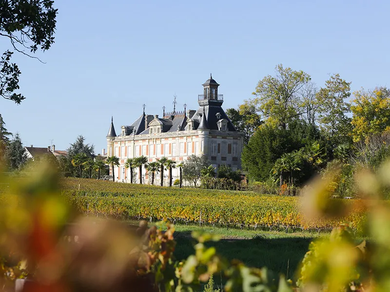 Marquis d'Alesme Becker Castle, a grand Bordeaux-style château with towers and mansard roof, surrounded by autumn vineyards in the Bordeaux wine region.