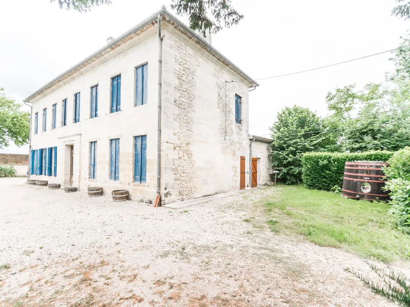 White stone house with blue shutters in a Bordeaux vineyard, featuring a gravel courtyard, wine barrel, and outbuildings.
