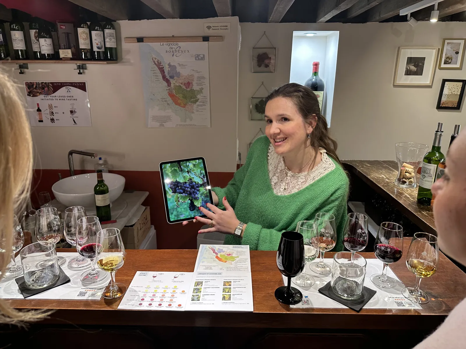 A smiling woman in a green sweater holds a tablet displaying an image of grapes during a wine tasting in Bordeaux, surrounded by glasses and wine information documents.
