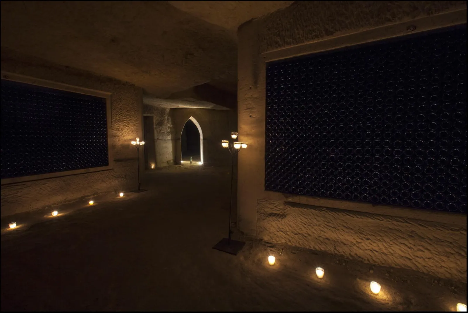 Underground gallery in Château de La Rivière, Bordeaux: stone-walled passage lit by warm golden lights, featuring dark blue textured panels and a gothic arch in the distance