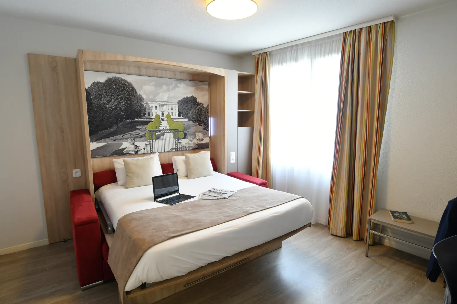 Modern hotel room with double bed, red and white pillows, architectural artwork above headboard, open laptop, large window with golden curtains, wooden flooring.