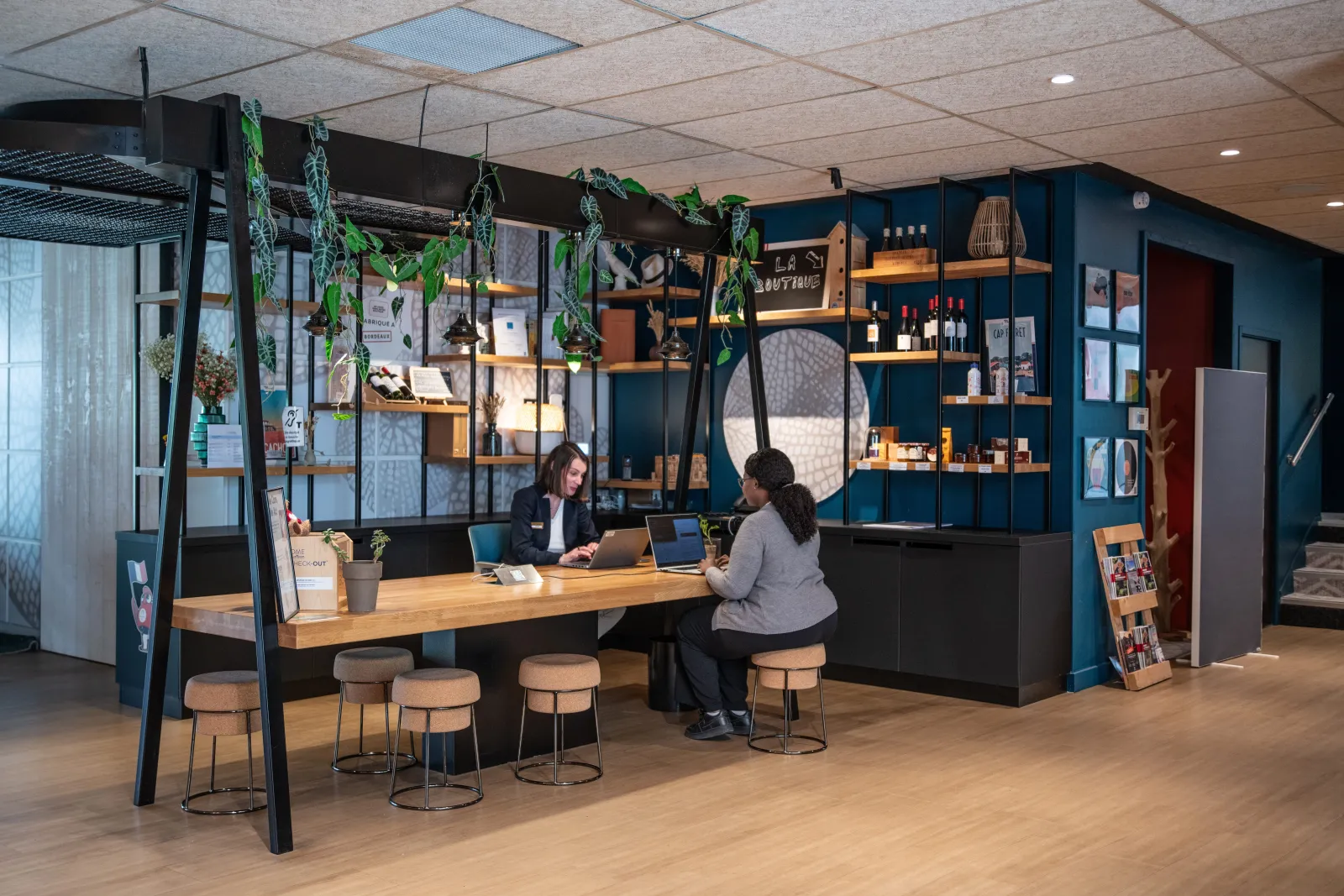 Modern reception area of Novotel Bordeaux Mérignac hotel featuring two staff members at a wooden desk, deep blue wall, illuminated shelving, hanging green plants, and beige bar stools.