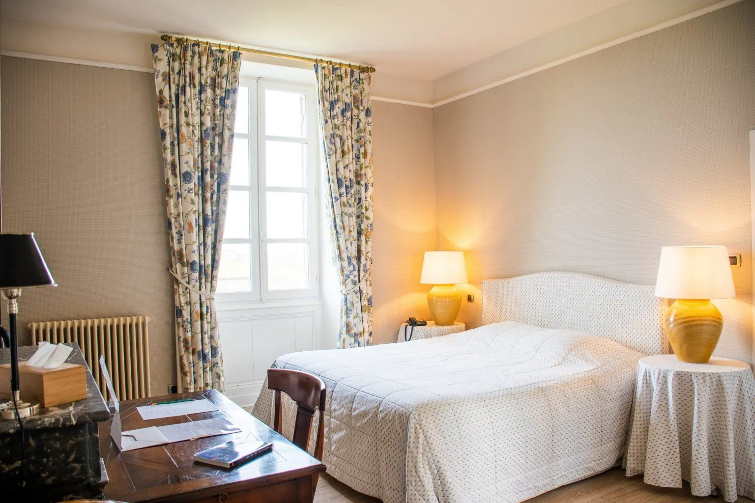 Elegant hotel bedroom featuring a double bed, golden lamps, floral patterned curtains, wooden desk, vintage radiator and street-facing window in Bordeaux.