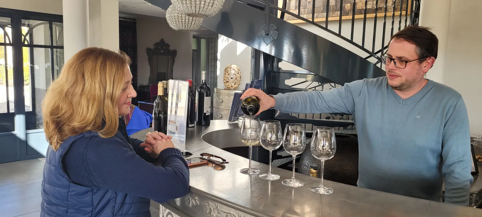 A sommelier pours red wine into a glass during a wine tasting at Château La Rose Perrière in Bordeaux, while a woman watches carefully.