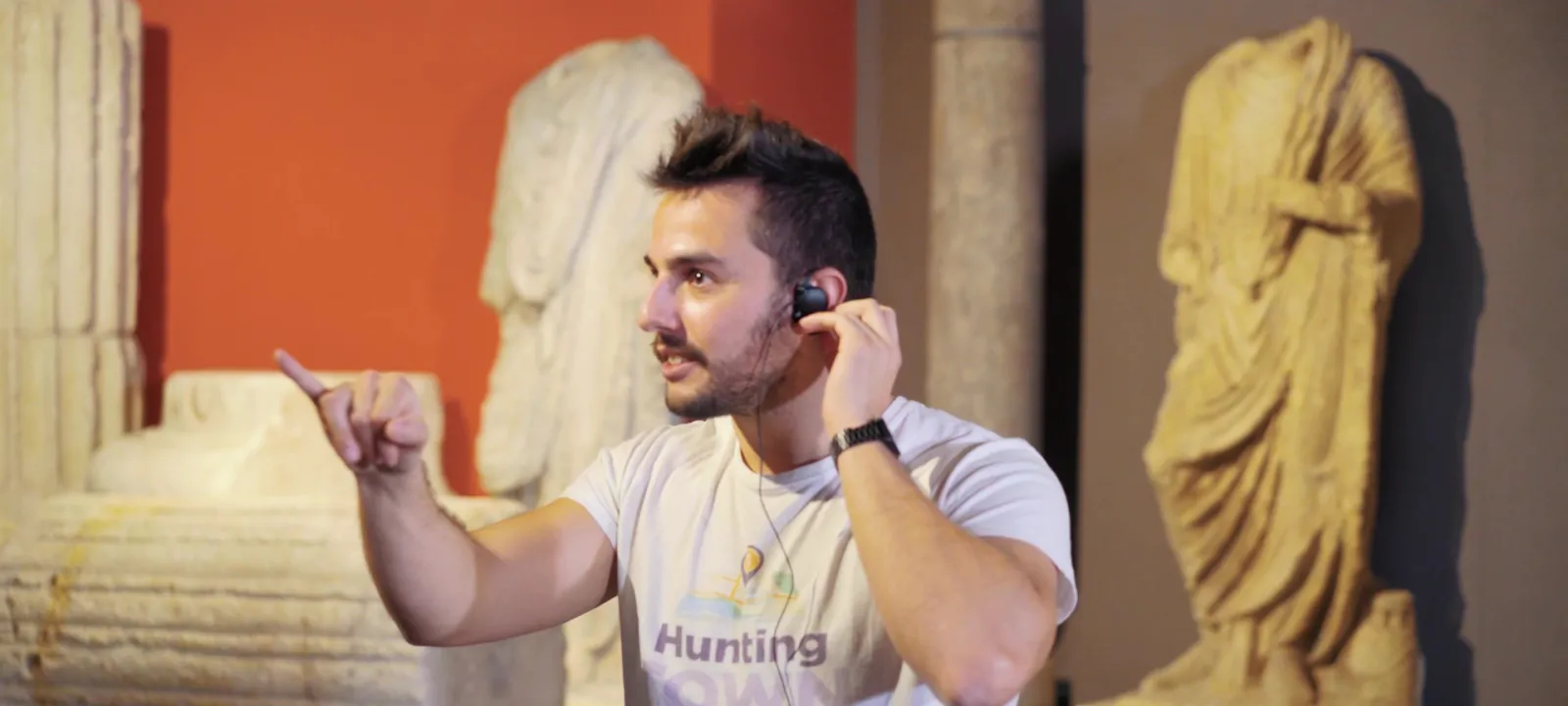 A man in a beige Hunting t-shirt talks on the phone in a museum exhibition, surrounded by ancient sculptures and orange walls.