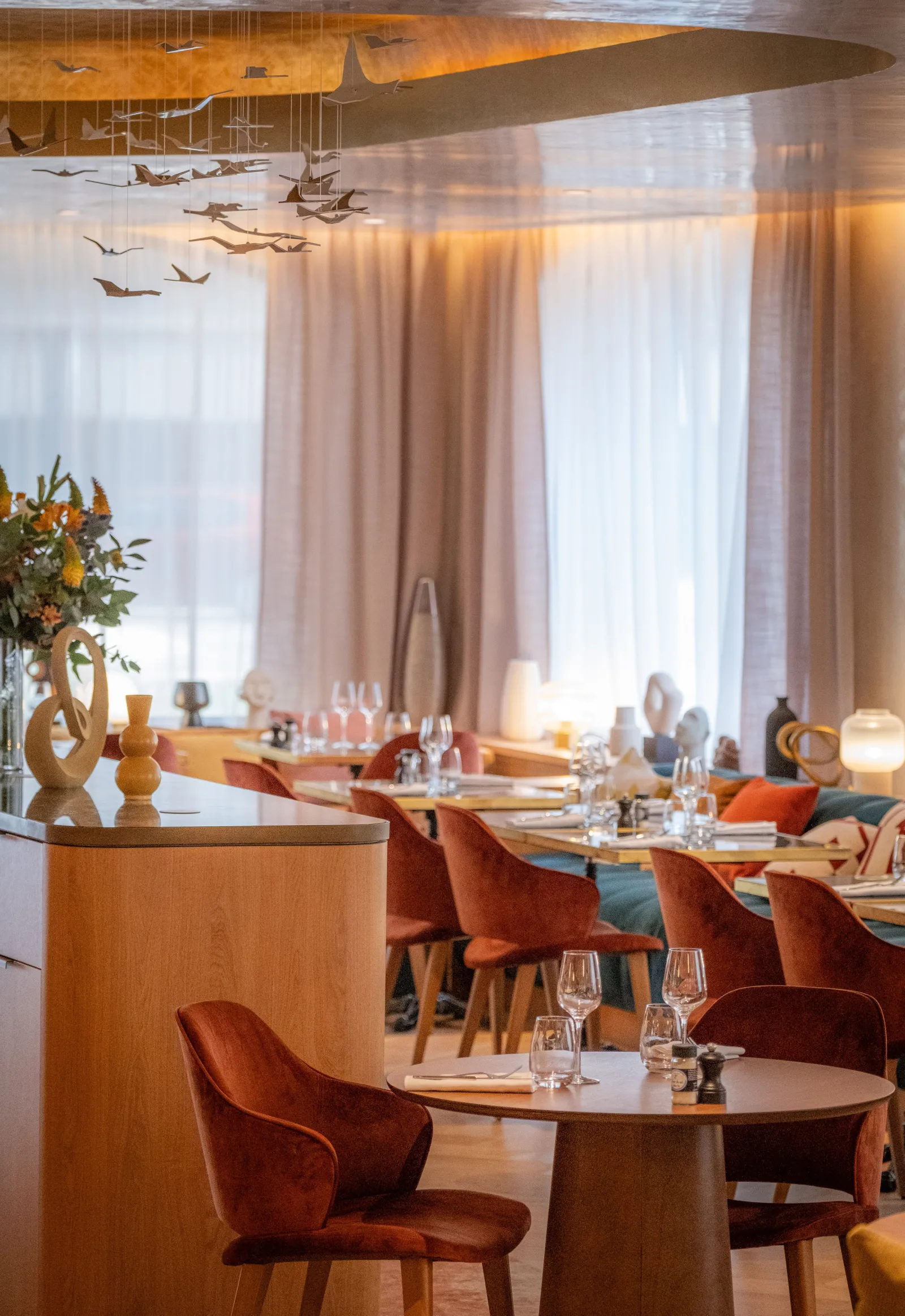 Elegant restaurant interior in Bordeaux featuring terracotta velvet chairs, round tables, large windows with beige curtains, natural wood décor, and an abstract ceiling sculpture.