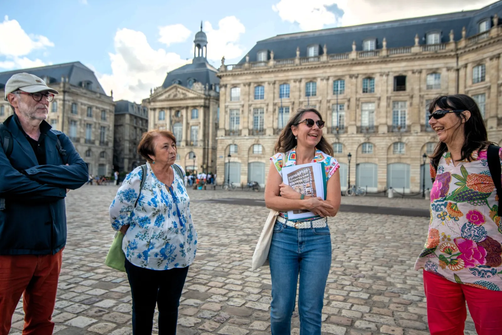 A group of tourists visit Place de la Bourse in Bordeaux, with historic stone buildings and classical architecture in the background.