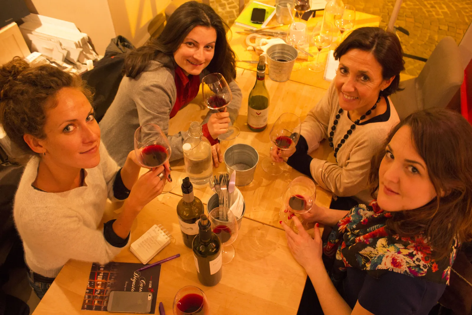 Five smiling women enjoying red wine tasting around a wooden table with wine bottles and glasses in a warm atmosphere in Bordeaux.