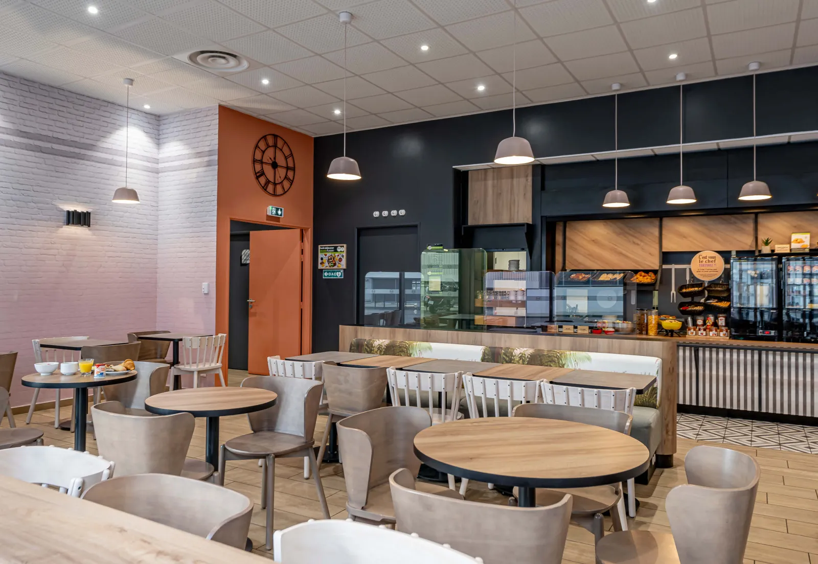 Modern café-hotel interior featuring round tables, beige chairs, service counter, orange and black walls, white coffered ceiling, and pendant lighting fixtures.