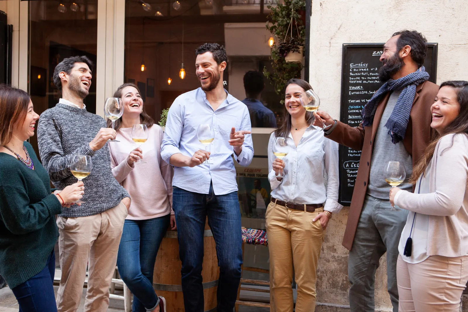 Group of seven people smiling and enjoying white wine outside a wine shop in Bordeaux, sharing a joyful evening atmosphere.