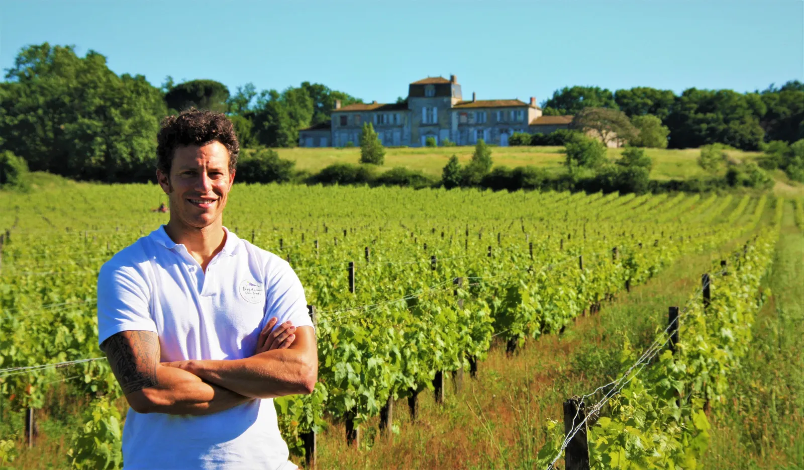 A smiling man in a white t-shirt standing in front of Bordeaux vineyards with a historic estate in the background