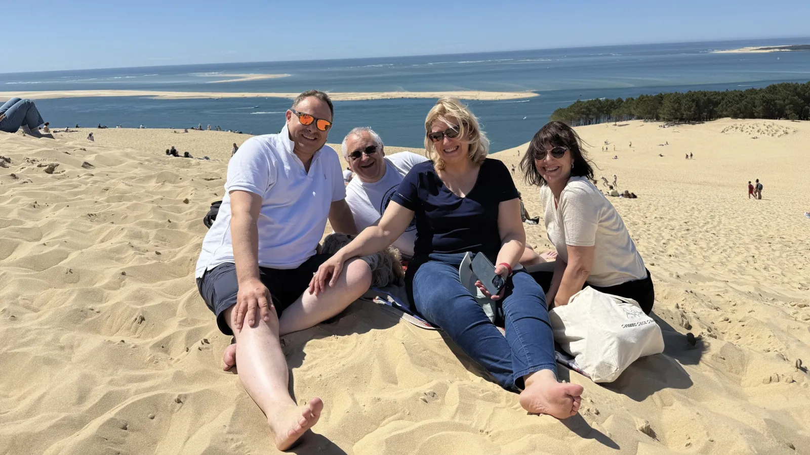Four smiling people pose together on a sandy beach with the ocean and forested dunes in the background.