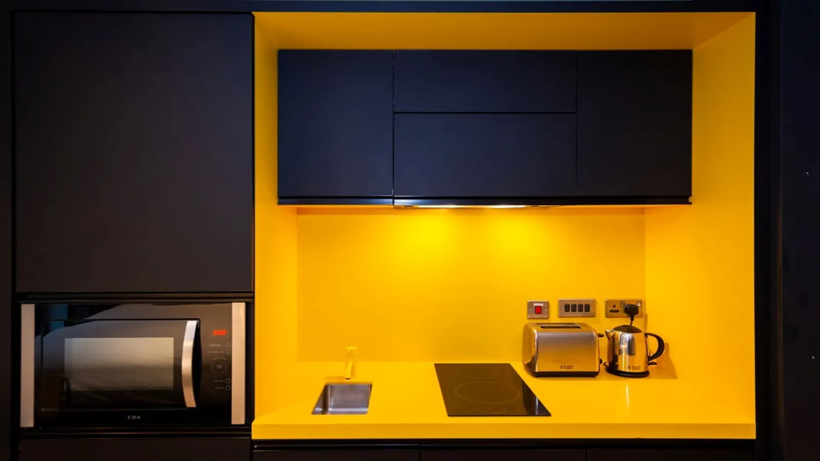 Modern kitchen in an aparthotel featuring bright yellow walls, wall-mounted flat-screen TV, built-in microwave, cooktop, toaster, kettle and electrical control panel