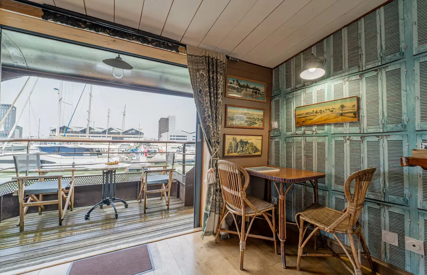 Interior of a barge with view of Bordeaux harbor, wicker furniture, framed paintings on walls and large windows overlooking moored boats