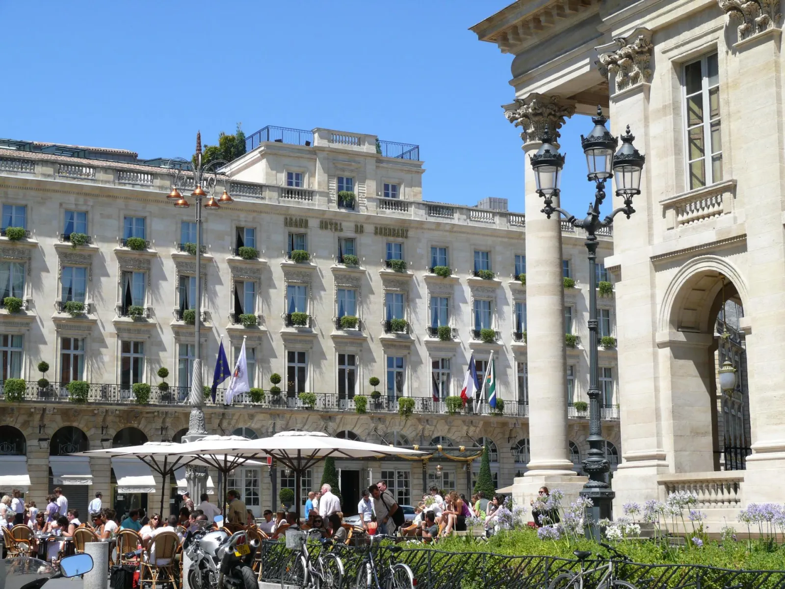 The Intercontinental Grand Hotel Bordeaux facade featuring its lively outdoor terrace, ornate balconies, Corinthian columns and flags under clear blue sky.