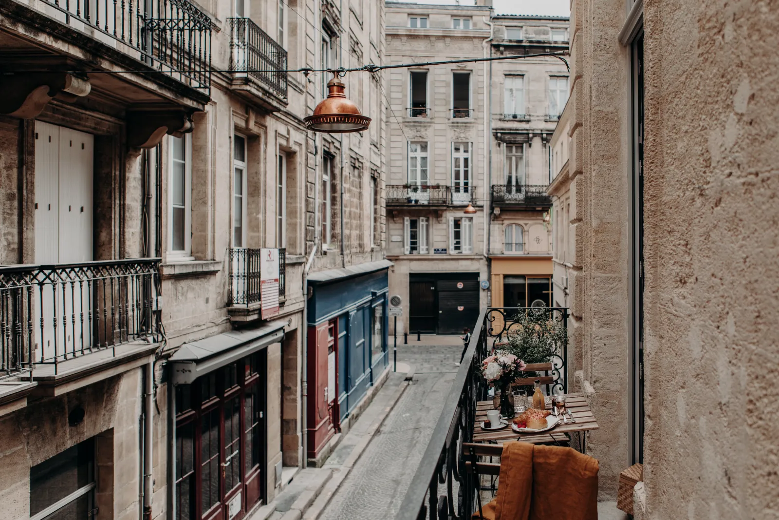 Narrow Parisian-style alleyway in Bordeaux featuring Haussmann buildings, a small decorated terrace with flowers and seating on the right side, and a vintage hanging lamp