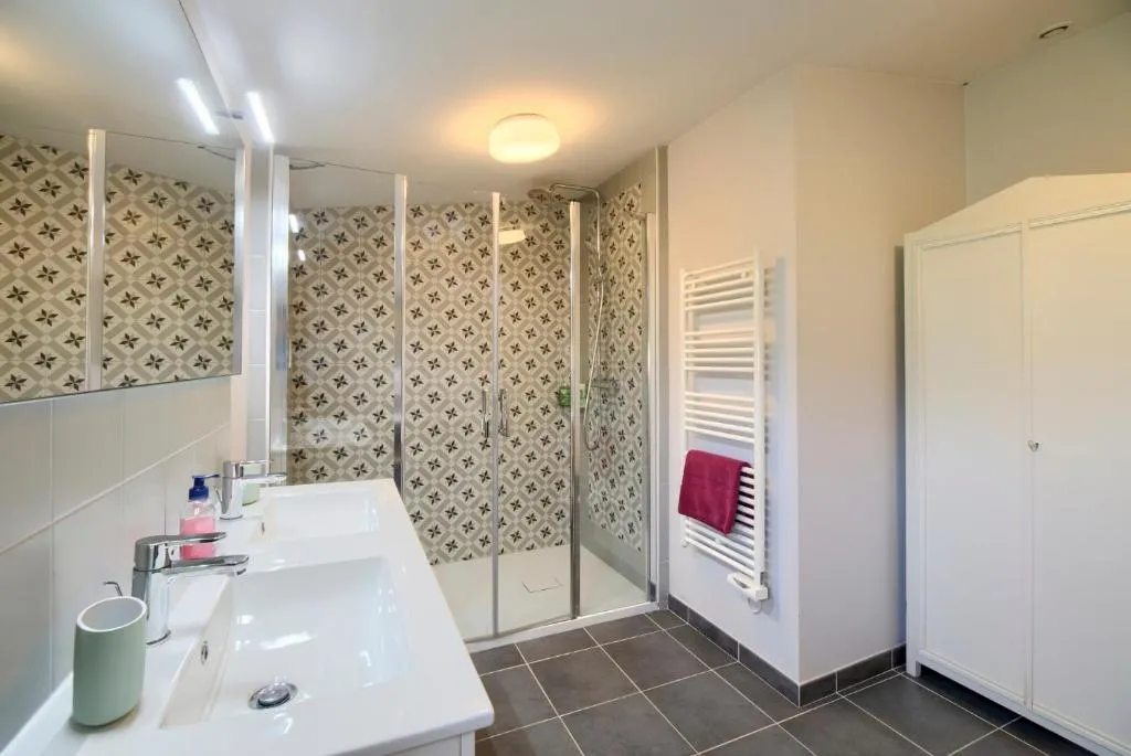 Modern bathroom featuring dual sink vanity, mirror, walk-in shower with glass enclosure, dark gray tiles, geometric patterned beige wallpaper, white towel radiator, and bathroom accessories.