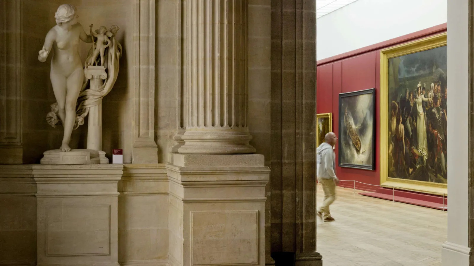 Gallery of the Musée des Beaux-Arts in Bordeaux featuring a white marble classical statue on a pedestal, Corinthian columns, and old master paintings on red and yellow walls. A visitor views the artworks.
