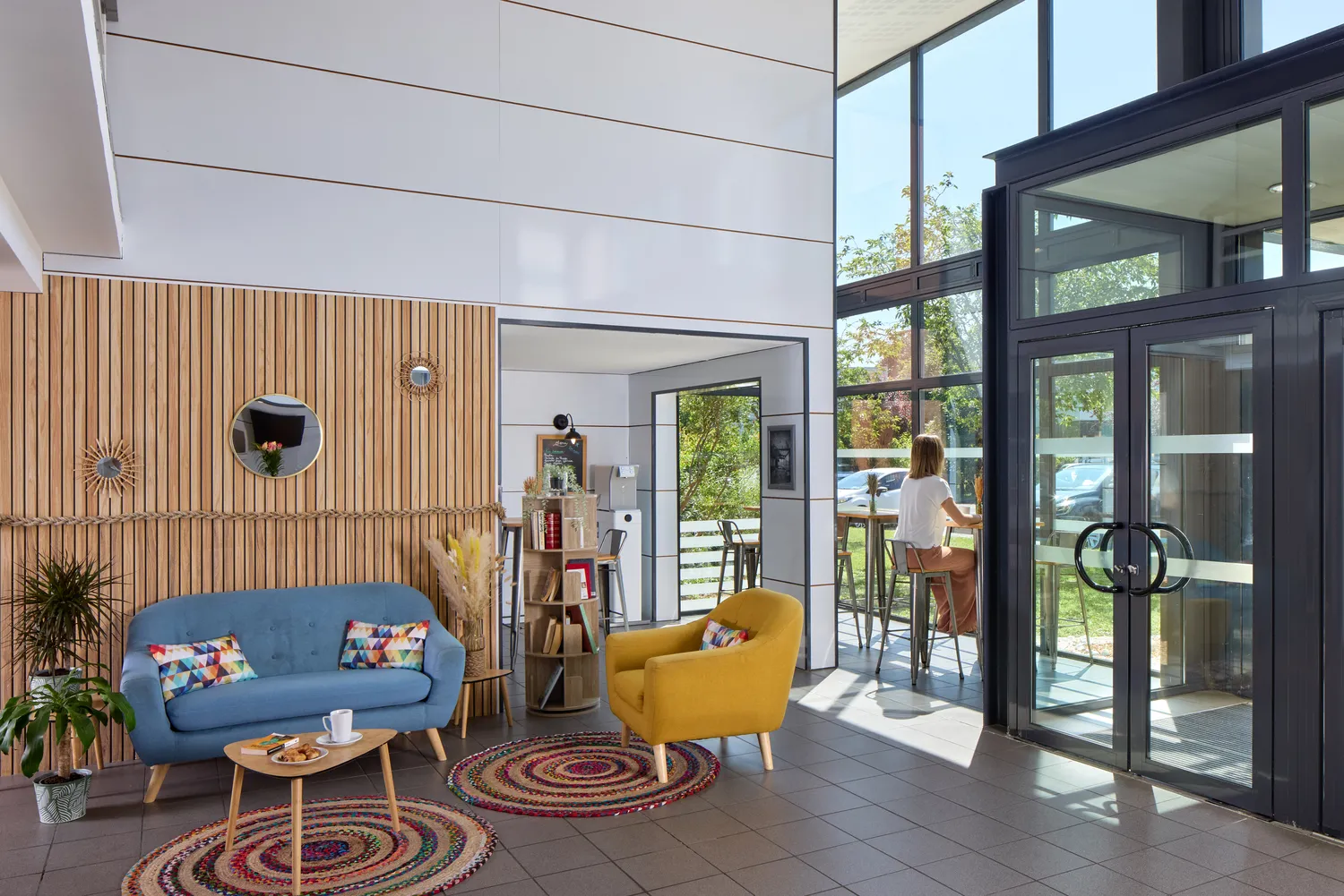 Bright modern living room featuring a blue sofa, yellow armchair, colorful rugs, and large glass doors opening onto a sunny patio, with light wood paneling and Scandinavian furniture.