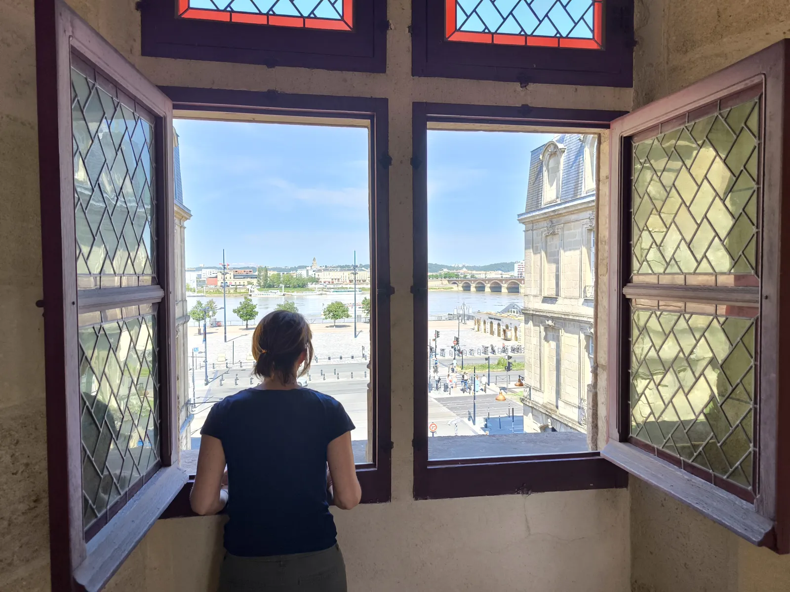 A woman seen from behind looking out through the open windows of Porte Cailhau in Bordeaux, overlooking the Garonne River, Pont de Pierre bridge, and Place de la Bourse square