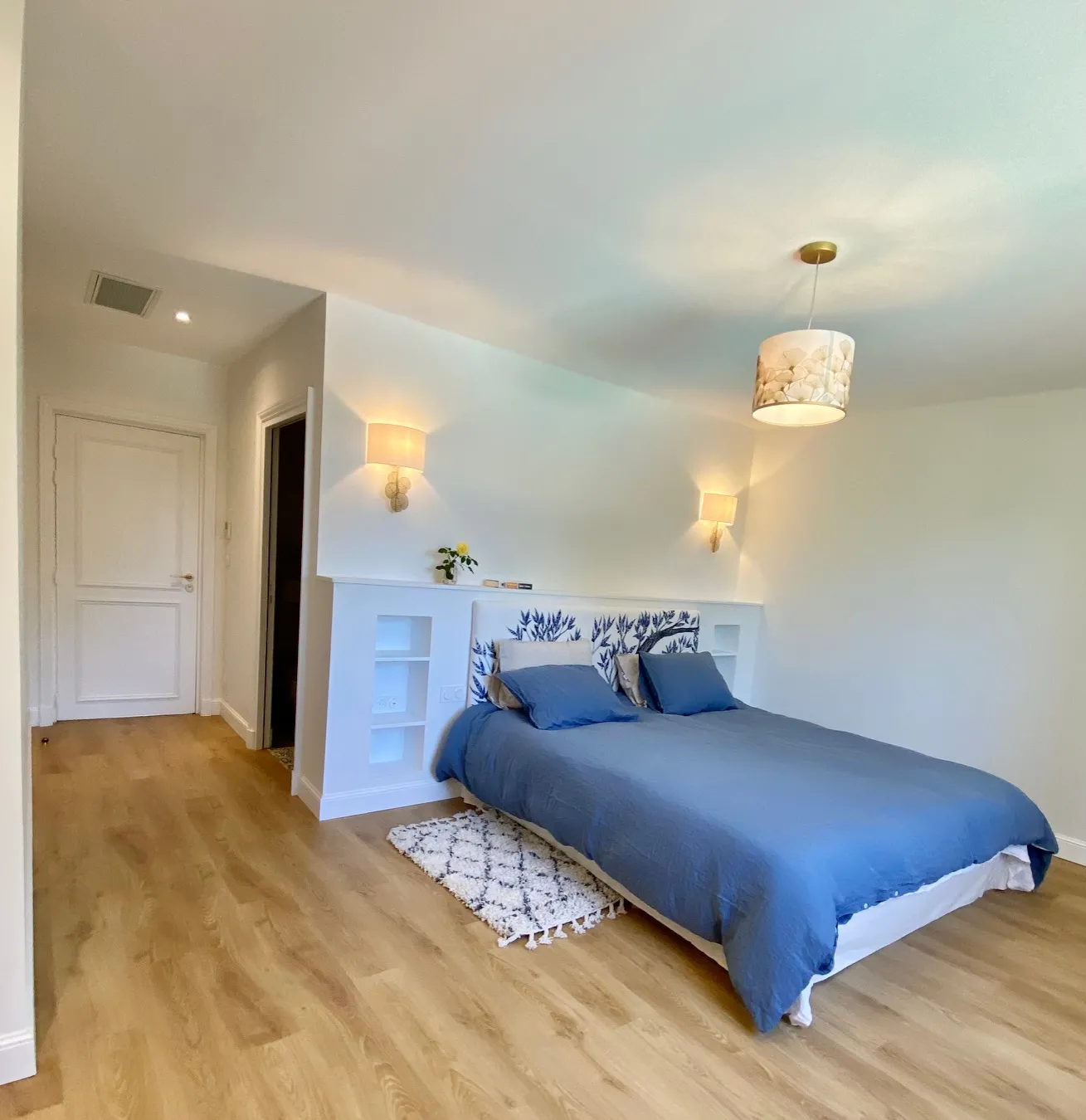 Modern bedroom featuring a queen-size bed with blue linens, floral-patterned headboard, light wood flooring, wall sconces and pendant lighting, built-in white storage cabinets, and entrance doors on the left