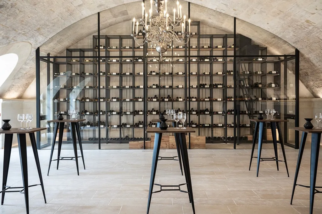Modern interior of a Bordeaux wine cellar featuring a large glass and metal display wall with numerous wine bottles beneath a stone vault. Black high tables with glasses are arranged throughout the space.