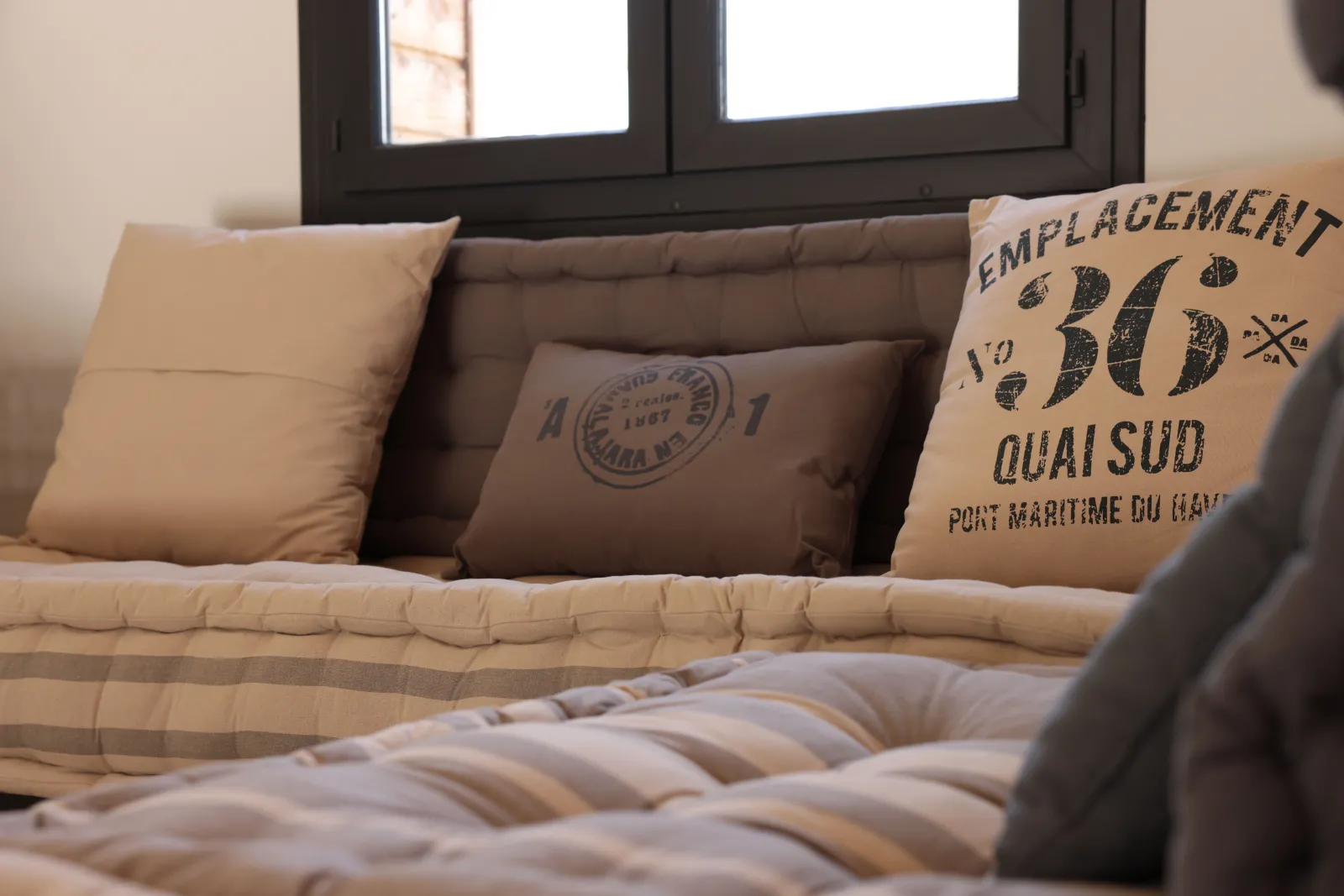A gray sofa with beige and brown pillows featuring maritime text, positioned in front of a black window at the Éco-Cottage Alfred in Bordeaux, Château Loudenne.
