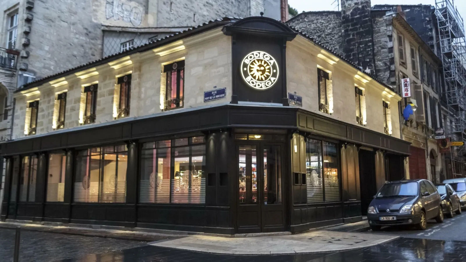 Illuminated facade of Bodega Bodega restaurant in Bordeaux, a corner building with clock, arched windows and dark glass storefront in the city center