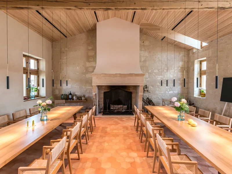 Elegant dining room with two long wooden tables, matching chairs, central stone fireplace, wooden ceiling, and side windows at Château Marquis d'Alesme in Bordeaux