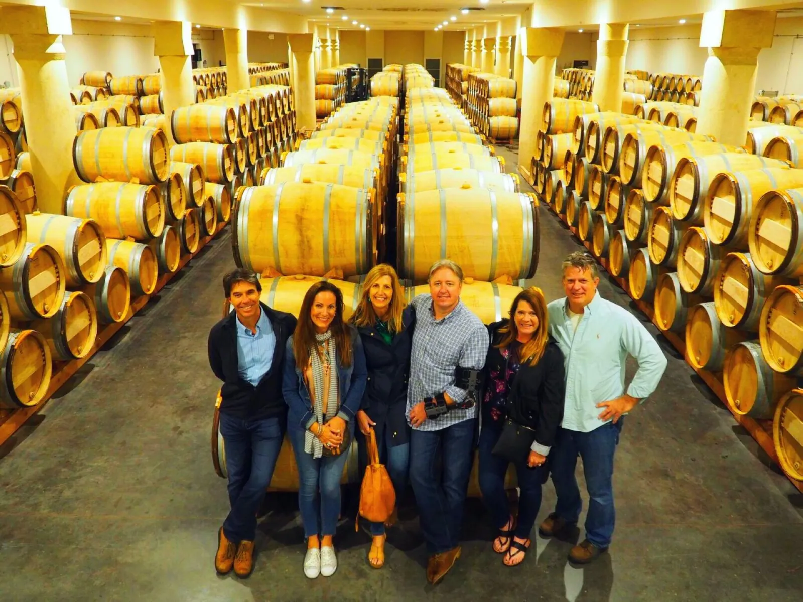 Six smiling people posing in a Bordeaux wine cellar lined with wooden barrels for wine aging.