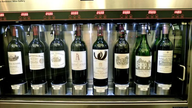 Refrigerated display case at Max Bordeaux Wine Gallery showcasing eight premium Bordeaux wine bottles with château labels and price tags displayed above each bottle.