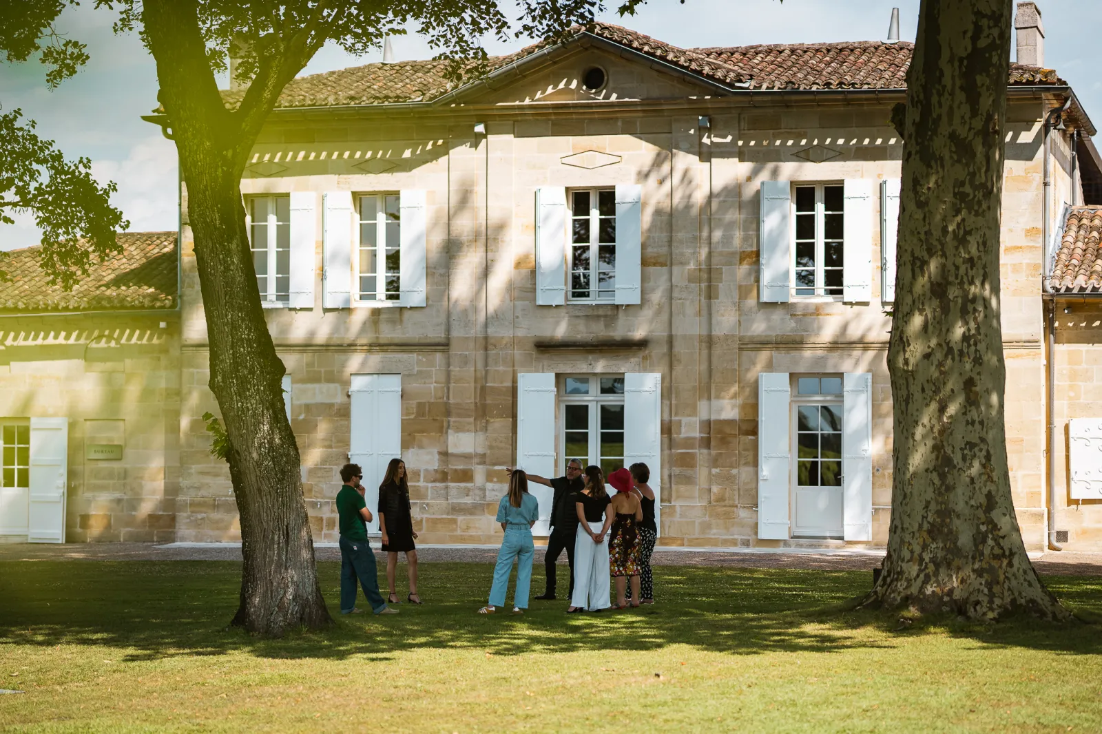 A group of six people standing beneath two large trees in front of an elegant white stone house with blue shutters in the grounds of Château La Dominique, Bordeaux.