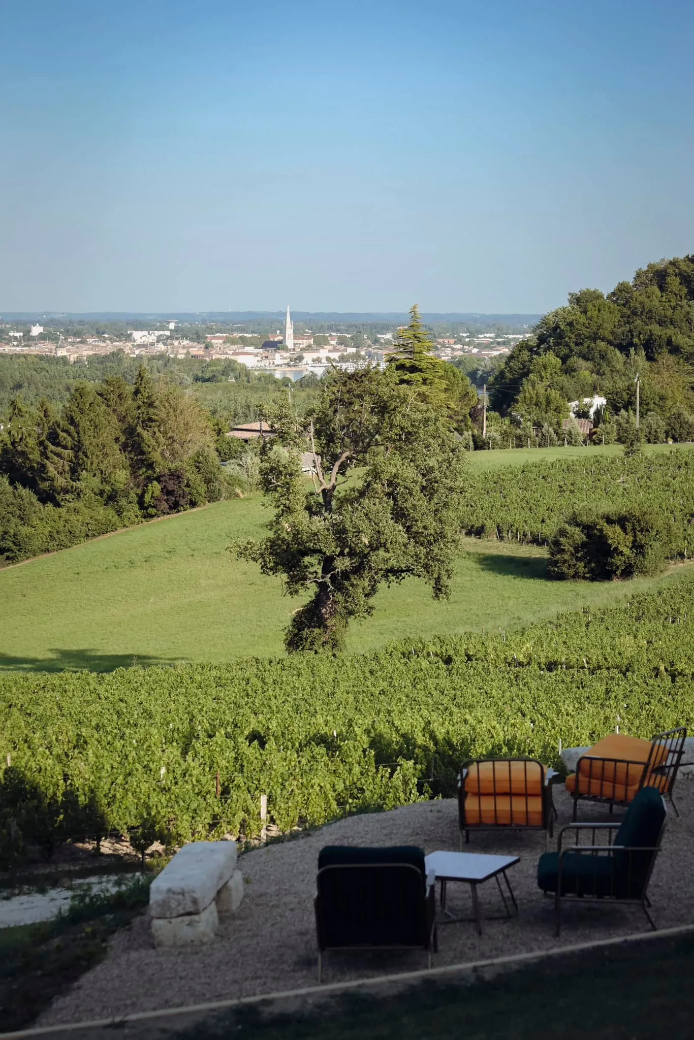 Panoramic view from a terrace with garden furniture overlooking rolling vineyards, with Bordeaux city and its church steeple in the distance under clear blue sky.
