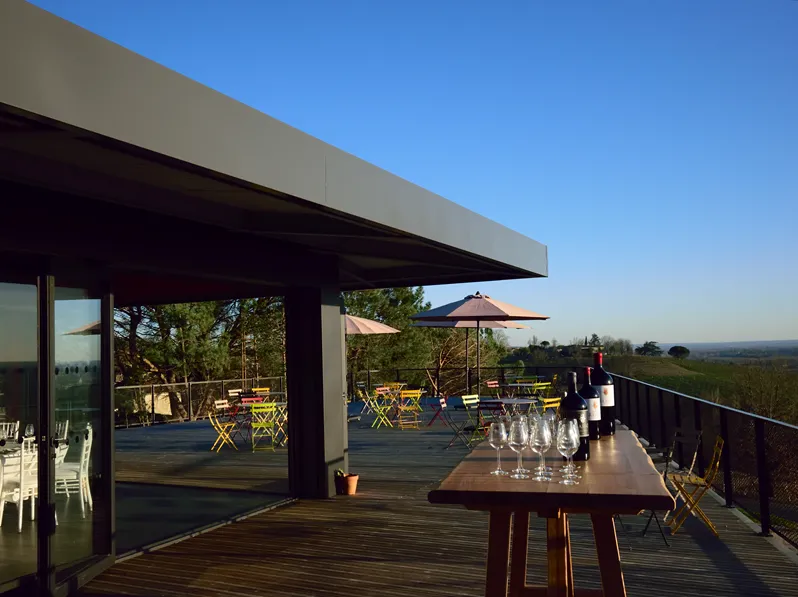 Modern terrace at Château Réaut near Bordeaux featuring high tables, yellow chairs, umbrellas, and panoramic views of the vineyards and countryside under clear blue skies.
