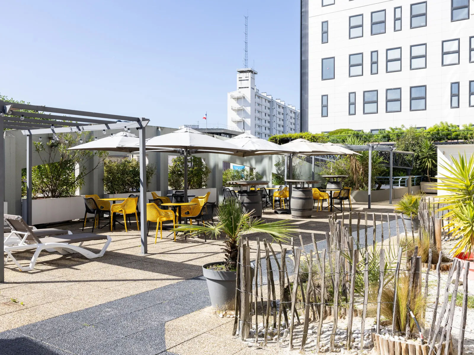 Sunny terrace of Bistrot de Mériadeck in Bordeaux featuring yellow chairs and tables under white pergolas with lush greenery and modern buildings.