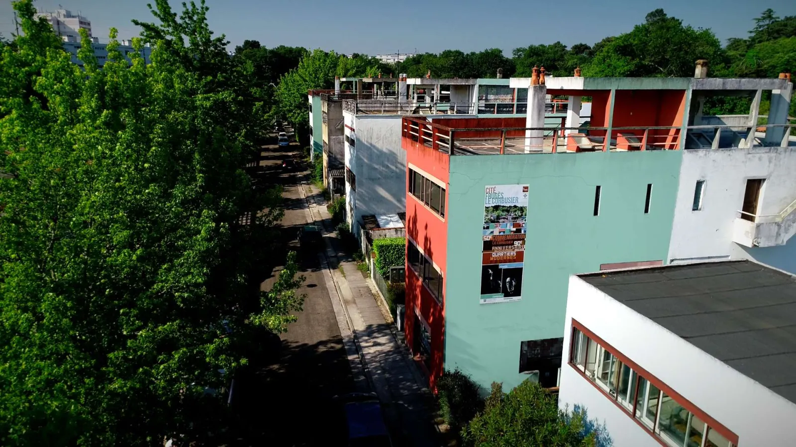 Aerial view of Cité Frugès in Pessac featuring colorful modernist buildings with turquoise and red facades, flat roofs, and terraces surrounded by lush greenery, designed by Le Corbusier.