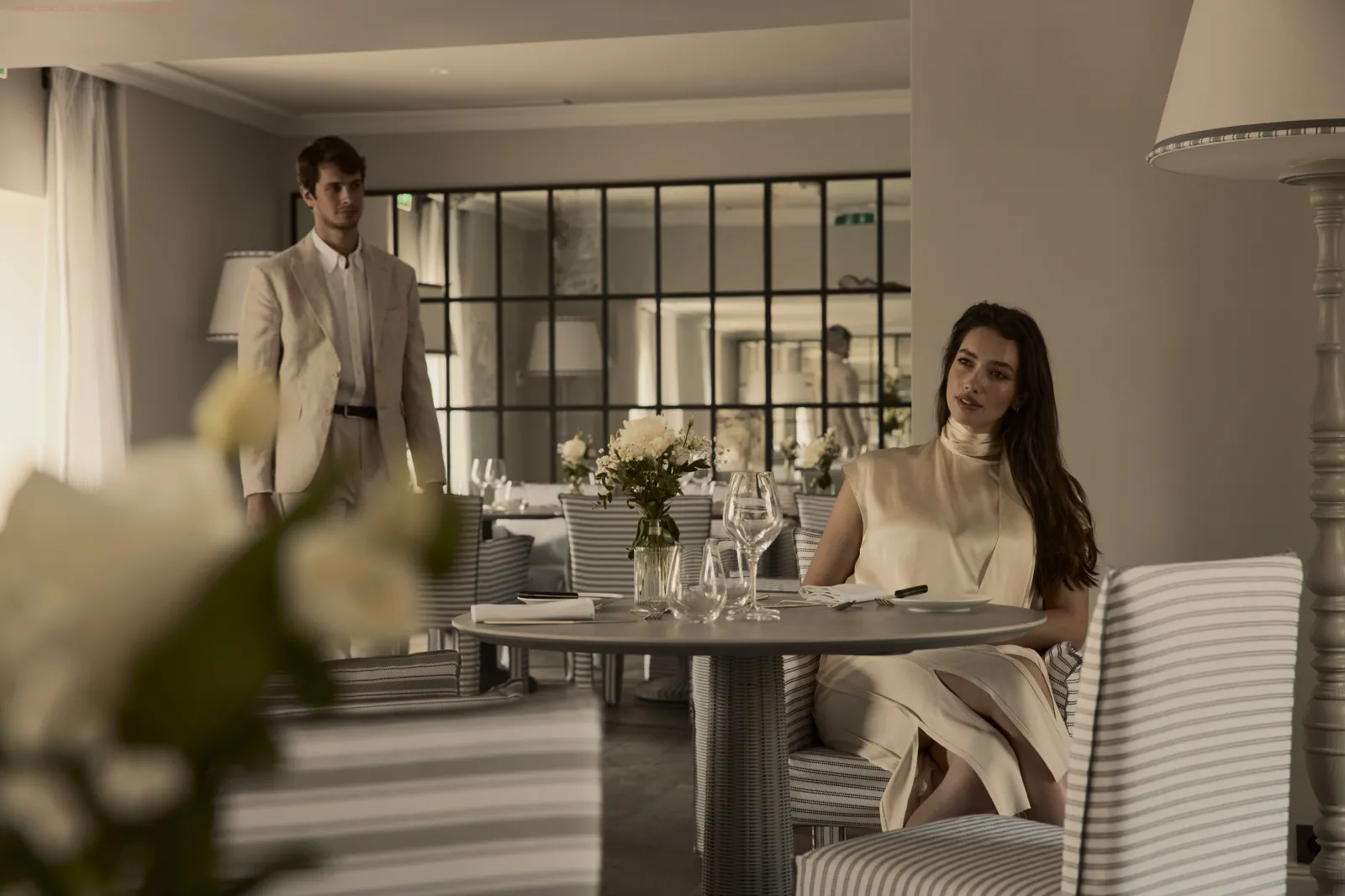 Two elegantly dressed people in white attire in an upscale restaurant featuring round tables, fresh flowers, and large grid windows overlooking the exterior.