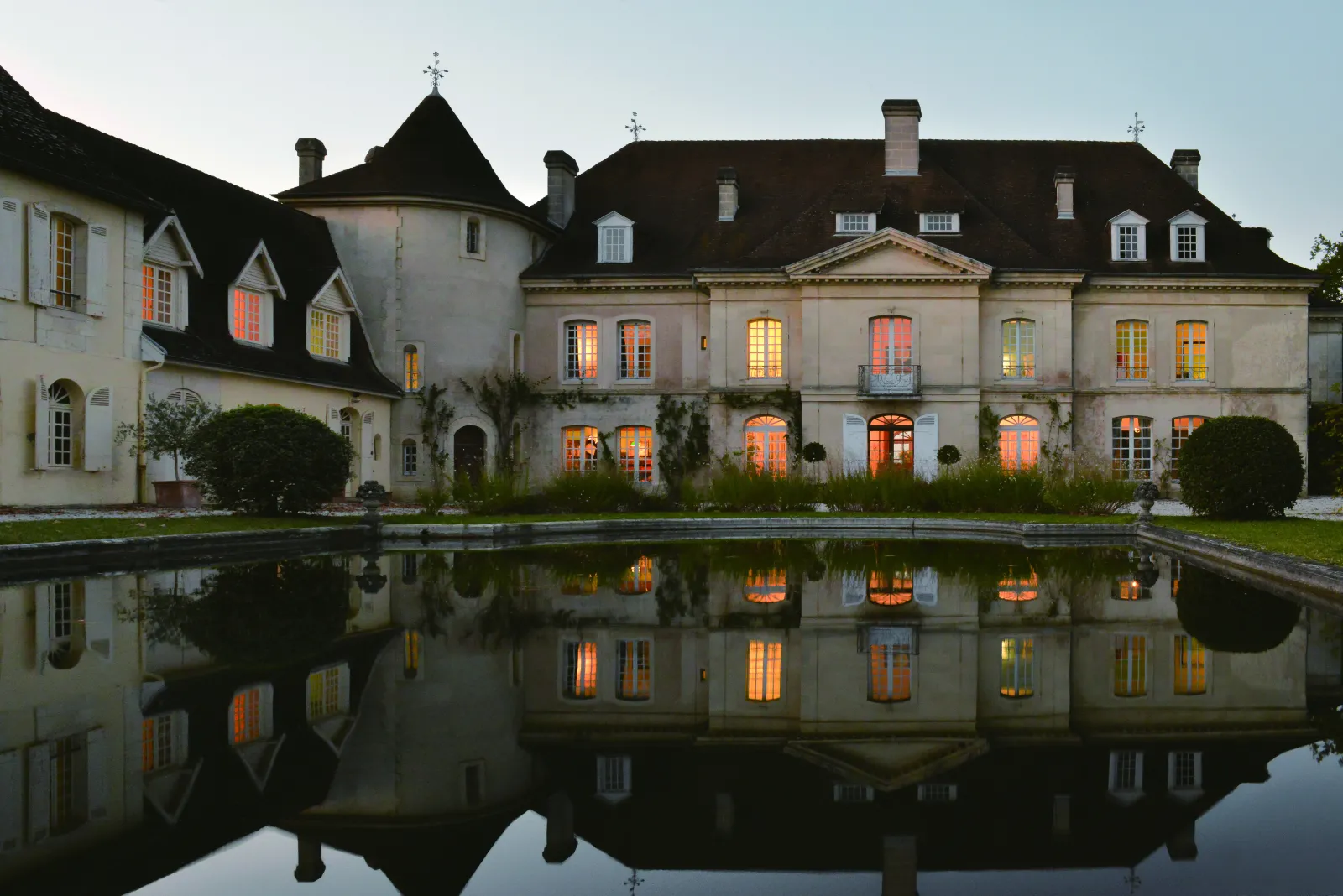Château Bouscaut in Bordeaux with illuminated facades mirrored in a water basin at dusk, featuring classical architecture with mansard roofs and ornamental crosses.