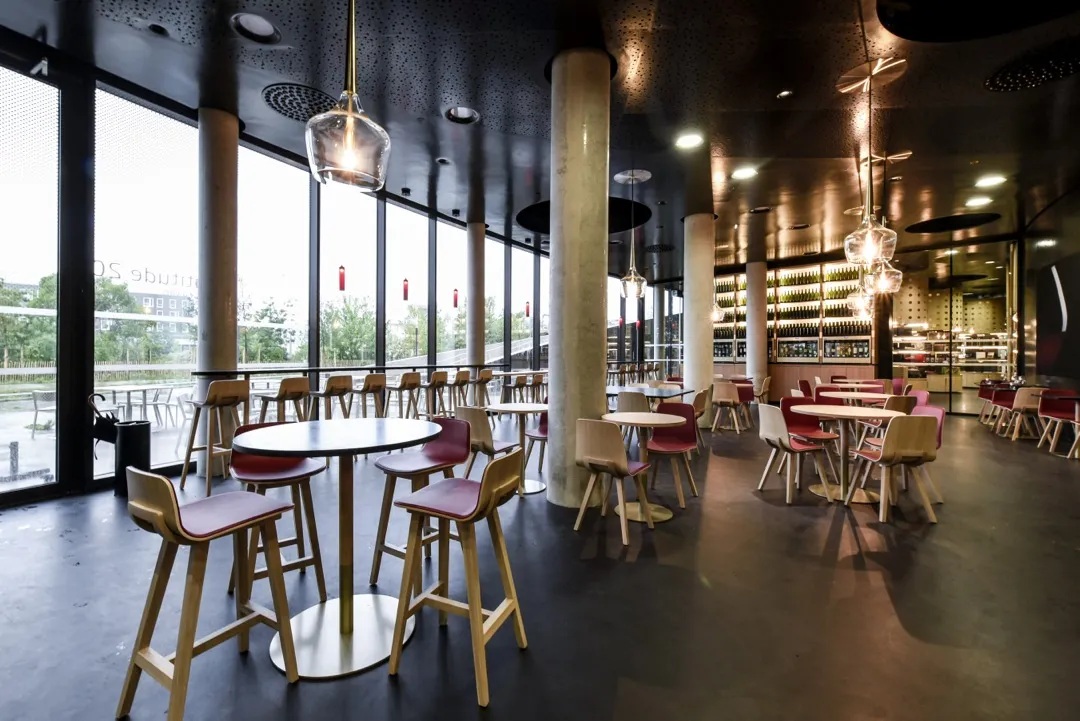 Modern interior of Latitude 20 wine bar in Bordeaux featuring high tables, colored chairs, concrete columns, large windows and warm lighting
