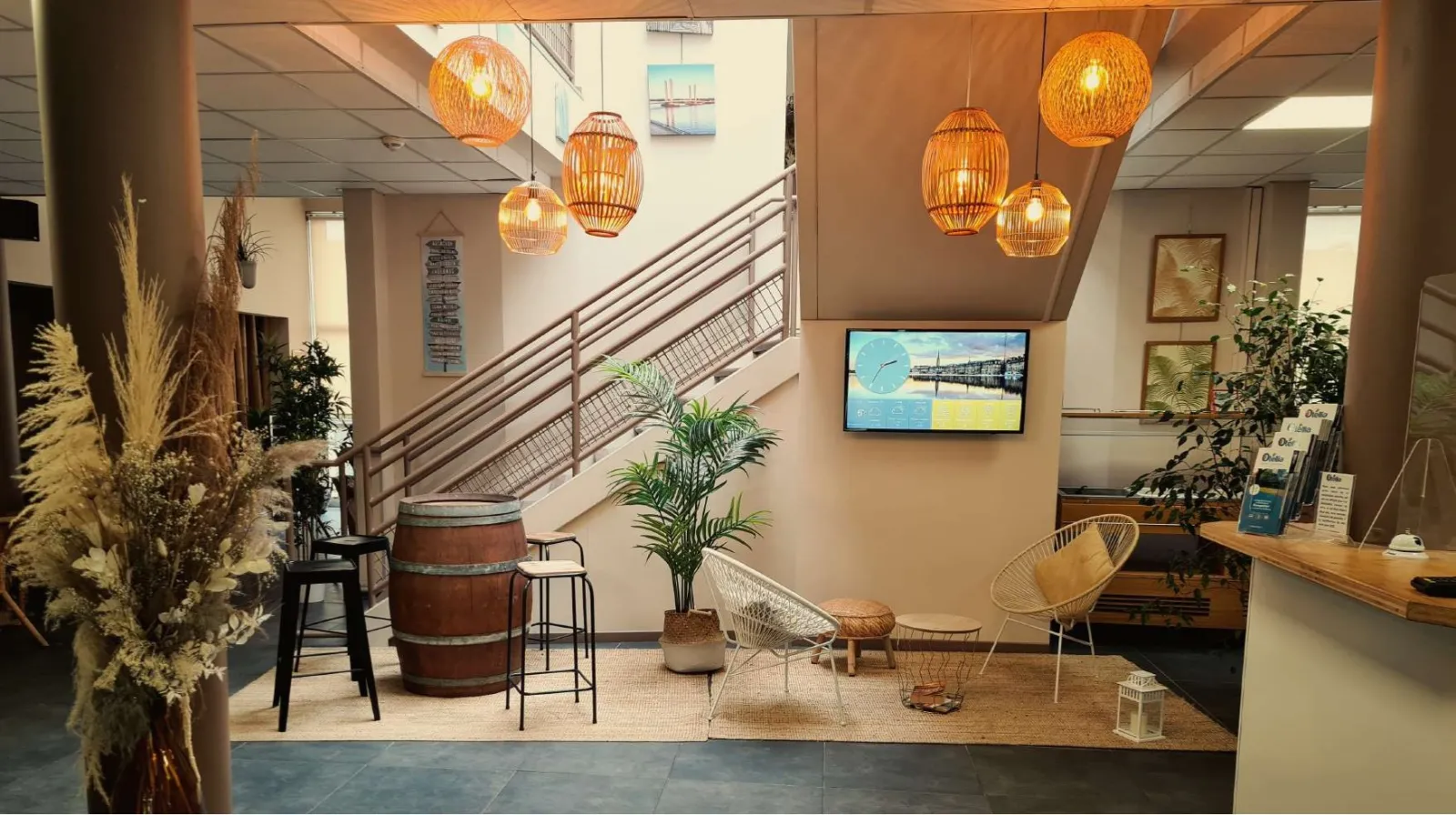 Modern lobby of Bordeaux Blanquefort Youth Hostel featuring a staircase, orange rattan pendant lights, designer furniture, decorative plants and relaxation areas.
