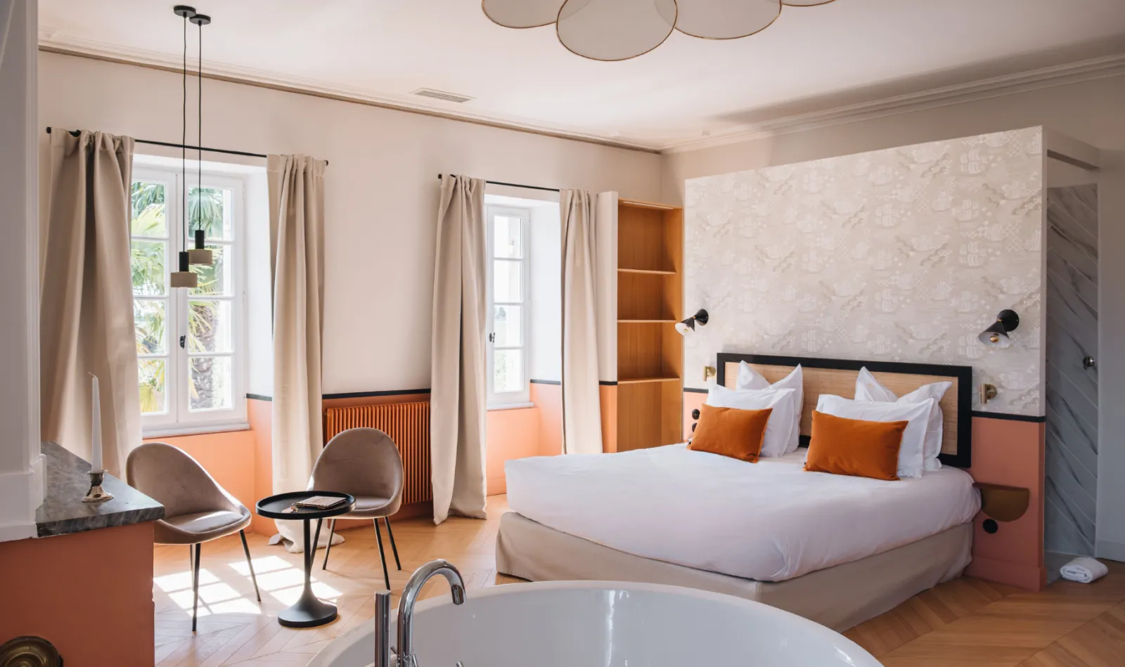 Luxury hotel bedroom featuring king-size bed, freestanding bathtub, modern furniture with orange and beige accents, large windows overlooking palm trees, at Château Réal in Bordeaux