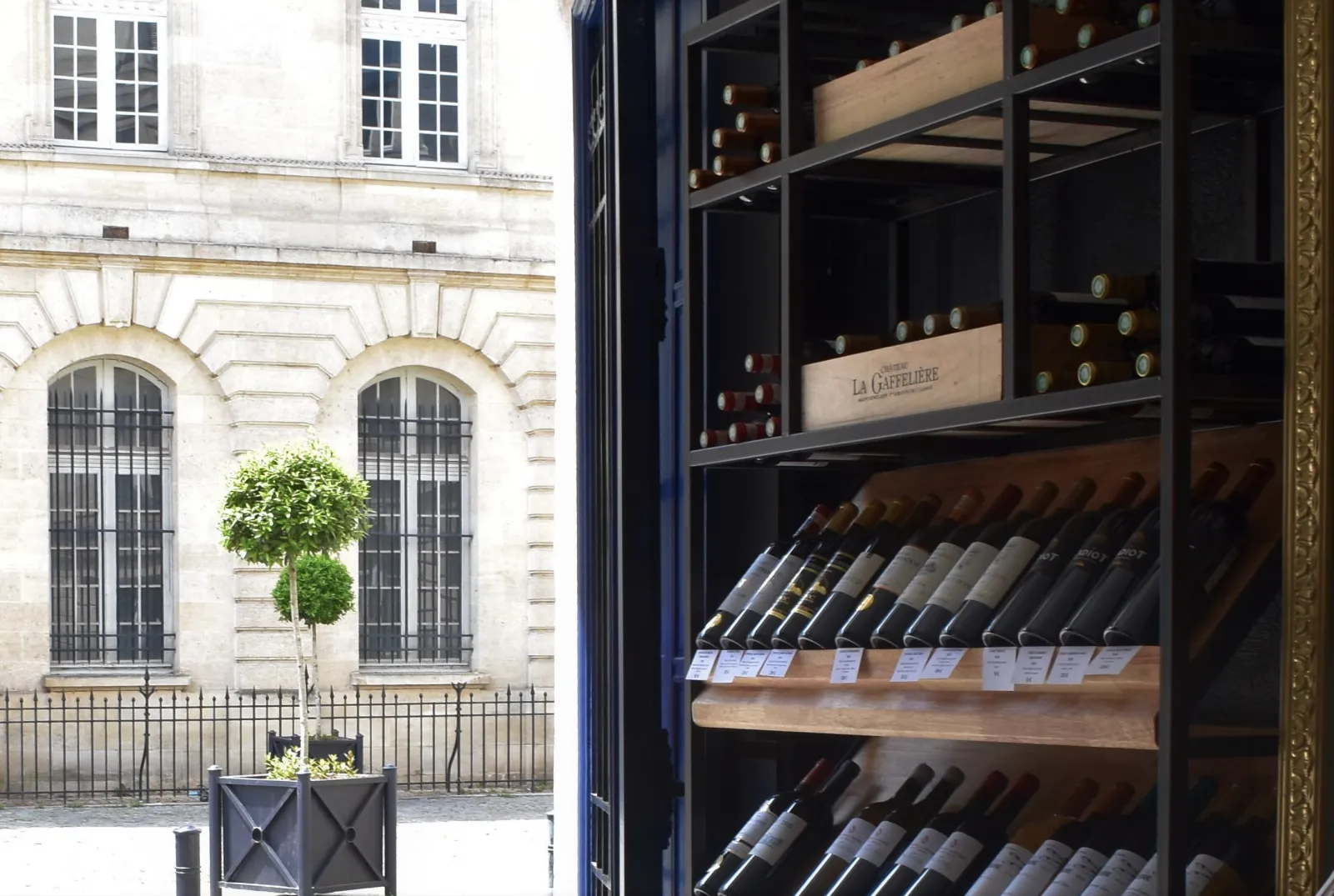 Wine shop storefront displaying wine bottles in dark glass cases, with a classic Haussmann building featuring arched doors and tall windows in the background.