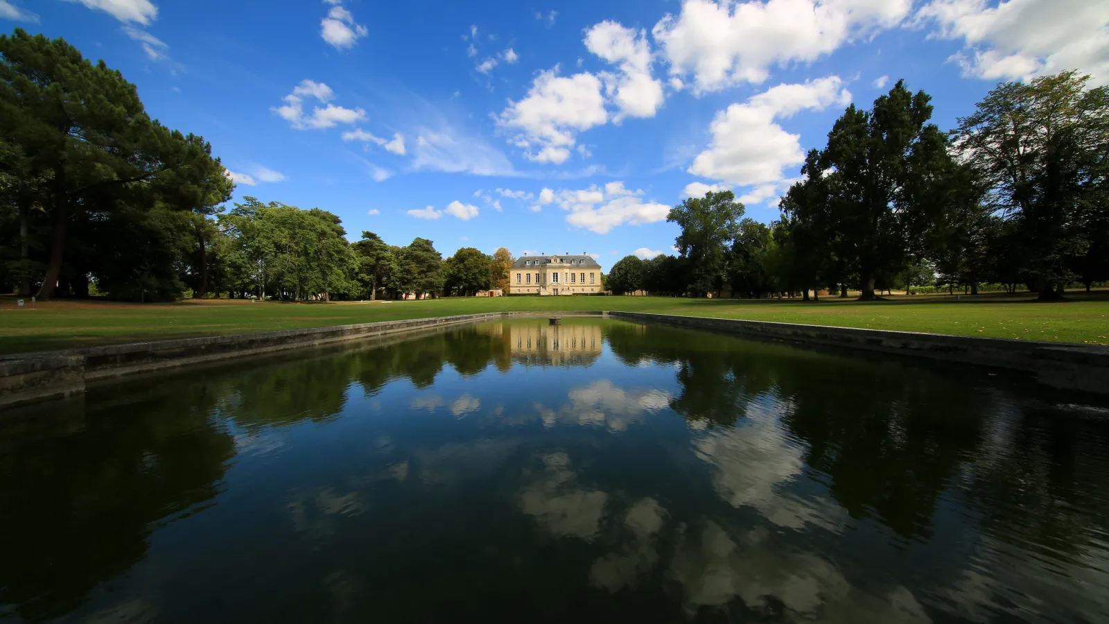 Château La Louvière near Bordeaux, a classical stone mansion with ochre-colored facade centered in a large manicured lawn, reflected in a canal with mature trees framing the estate under a clear blue sky.