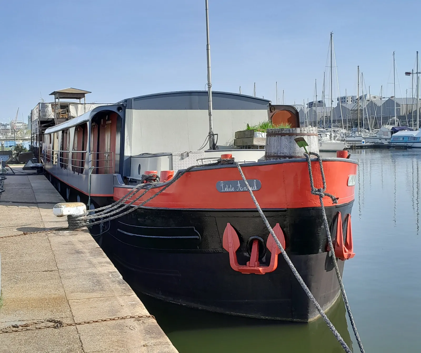 The Petite Annick houseboat moored at the quay in Bordeaux, featuring a red and black hull, surrounded by sailboats in the river port.
