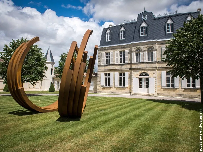 Abstract rust-colored metal sculpture in front of Château Malescasse in Bordeaux, featuring the castle's distinctive stone facade and mansard roof.