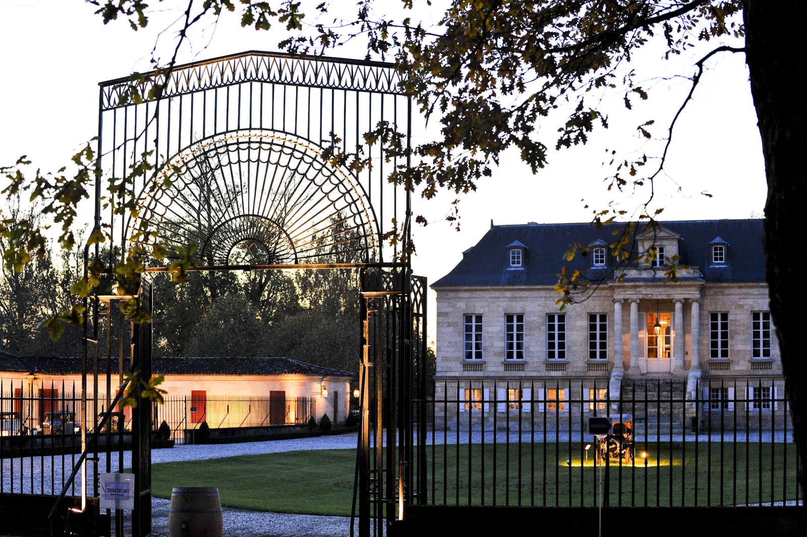 Ornate iron gate with Art Nouveau fan design at the entrance of Château La Louvière in Bordeaux, with the neoclassical mansion illuminated in the evening light.