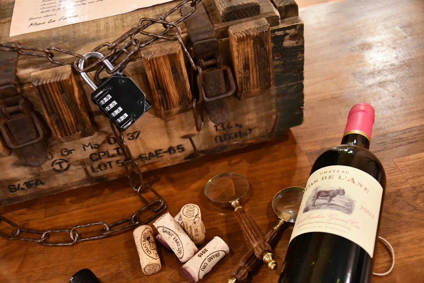 Wine-themed escape game scene featuring a wooden crate secured with chain and combination padlock, a Bordeaux wine bottle, cork stoppers, and tasting accessories on a wooden table.